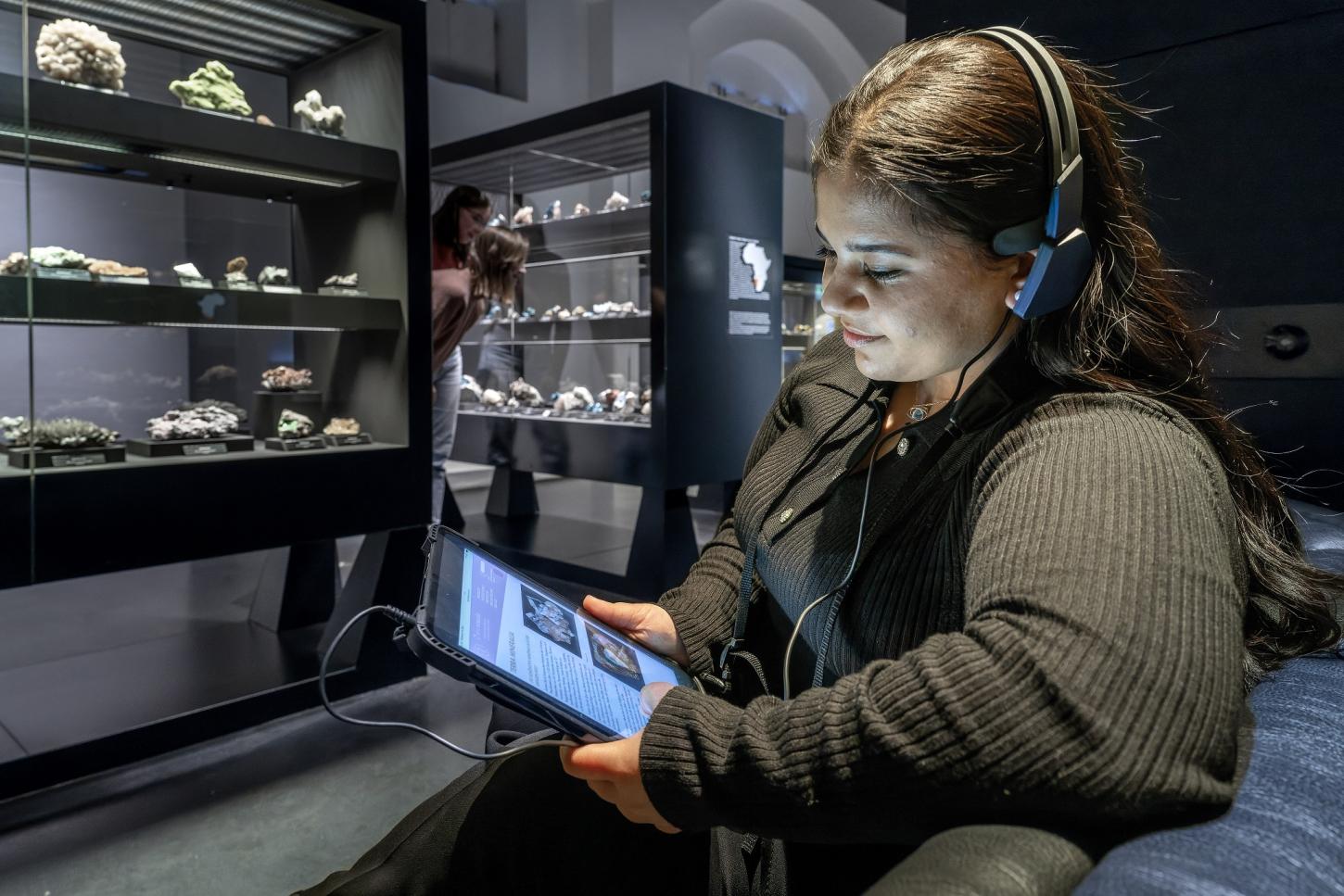 Eine junge Besucherin sitzt in der Ausstellung. Sie trägt Kopfhörer und hält ein Tablet, um sich den Audio-Guide anzuhören.