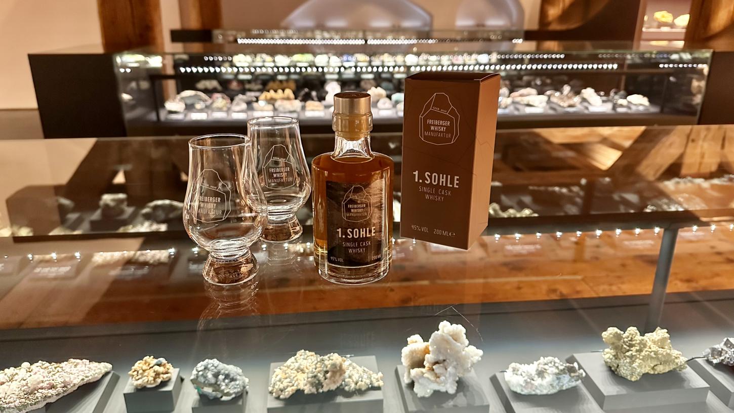 Eine braune Whiskyflasche, zwei Gläser und eine Verpackung stehen auf einer Vitrine, in der sich Minerale befinden.