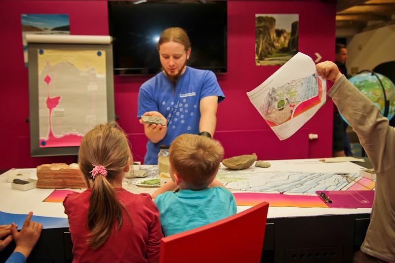 Ein Student im blauen T-Shirt zeigt zwei Kindern wie und wo Gesteine entstehen. Er hält ein Gestein in der Hand.